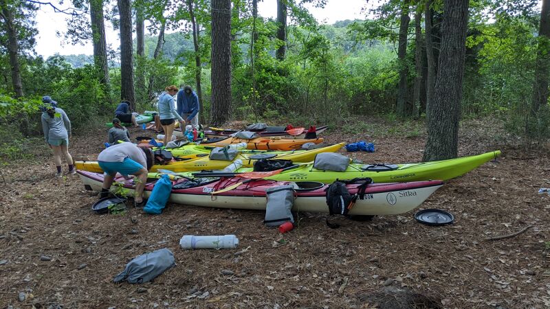A group of people are gathered around several kayaks on a wooded shore. The kayaks are various colors, including yellow, green, and red, and are loaded with gear. The people appear to be preparing for or returning from a kayaking trip. The ground is covered with leaves and pine needles, and trees surround the area.